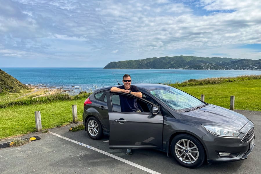 A driver stands next to their open door of their Wellington car rental parked by the coast not far from the city.