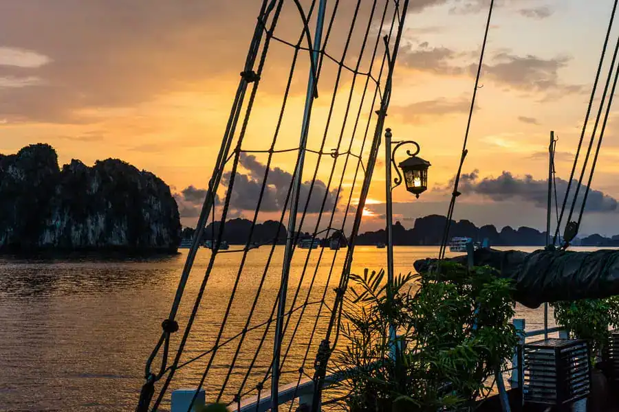 Looking past rigging to karst islands and a cloudy sky turning orange - cruise Bai Tu Long Bay and end your days like this.