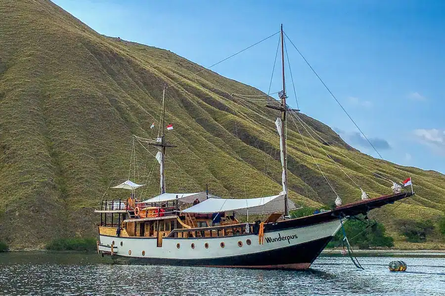 A wooden phinisi boat named Wunderpus anchored just off a sloping green hill while diving Komodo National Park.