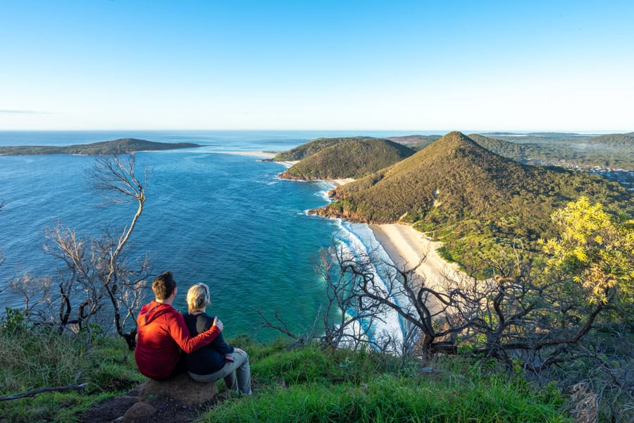 Two people travelling as a couple sit on a hill overlooking the sea and a beach.