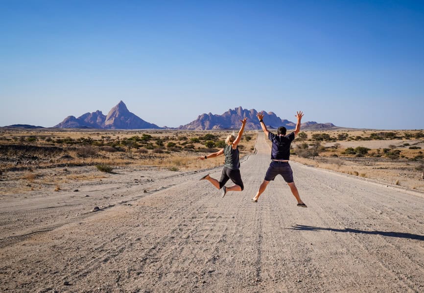 Two people jump into the air while travelling together on a long road with mountains in the distance.