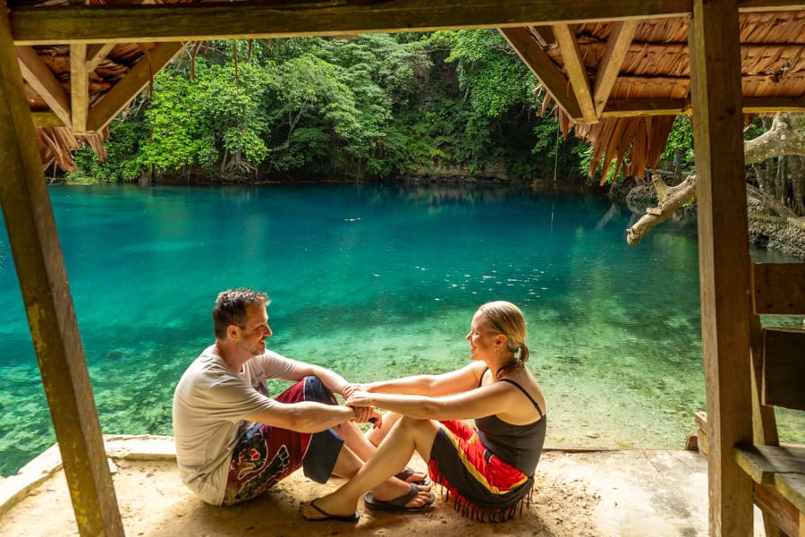 A traveling couple sit facing each other holding hands by a body of water.