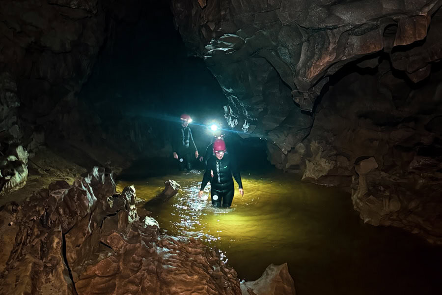 People in wetsuits with head torches walk through water in a cave – a unique adventure for couples who travel together.