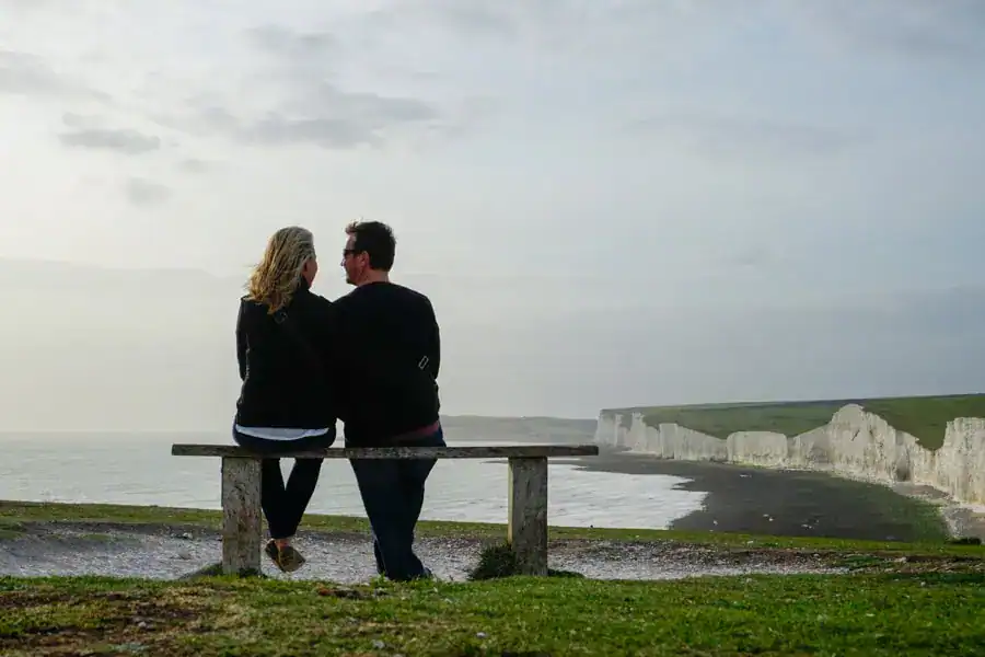 A travel couple sit together on a bench overlooking white cliffs and sea.