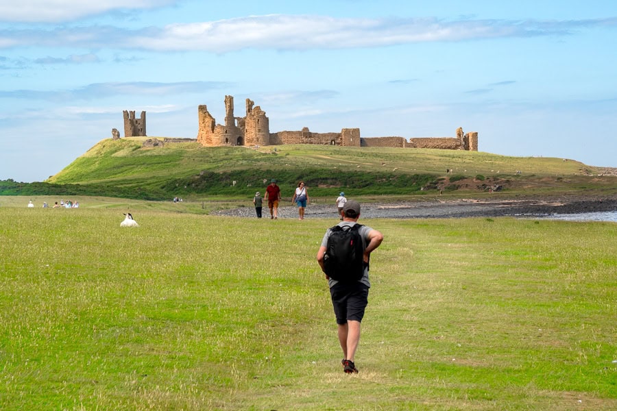 A man walks across coastal farmland towards the ruin of Dunstanburgh Castle, one of the best things to see in Northumberland.