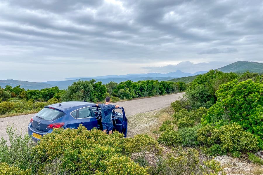 The driver of a blue car stands, with the door open looking across the hilly countryside towards the Costa Verde coastline.