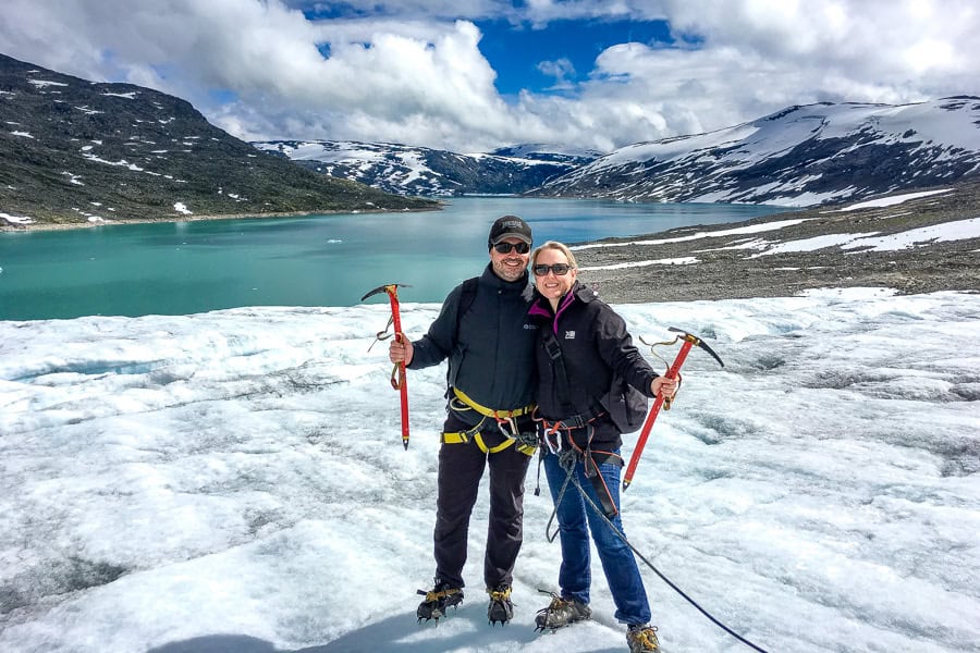 A couple decked out in glacier hiking gear stand on the ice with sundrenched Styggevatnet glacier lake in the background.